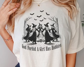 Puede incluir: Camiseta color crema con una ilustración en blanco y negro de cinco brujas en círculo alrededor de una hoguera, con murciélagos volando encima. El texto "God Forbid A Girl Has Hobbies" está impreso debajo.