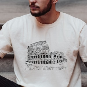 May include: A cream-colored t-shirt with a black and white illustration of the Colosseum and the text "Roman Empire on the Brain".