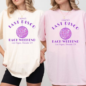 Op de afbeelding: Twee witte en roze t-shirts met de tekst "Laura's Last Disco Bach Weekend Las Vegas, Nevada '24" en een paarse discobal-afbeelding.