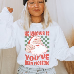 Op de afbeelding: Witte trui met een rode en groene afbeelding van de kerstman met de tekst "He knows if you've been flossing".