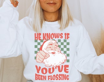 May include: White sweatshirt with a red and green graphic of Santa Claus with the text "He knows if you've been flossing".