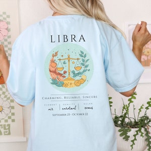 May include: Light blue t-shirt with a graphic of the Libra zodiac sign, featuring a scale, leaves, and citrus fruits. The text "LIBRA", "CHARMING, RELIABLE, SINCERE", "ELEMENT air", "QUALITY cardinal", "RULER venus", and "SEPTEMBER 23 - OCTOBER 22" is printed on the shirt.