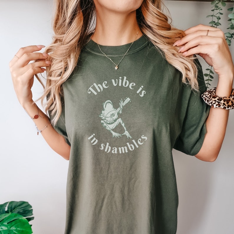Op de afbeelding: Olijfgroen T-shirt met de tekst "The vibe is in shambles" en een lichtgroene afbeelding van een kikker die een snaarinstrument bespeelt. De tekst en afbeelding staan in het midden.
