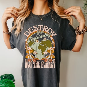 Puede incluir: Una camiseta gris oscuro con un dise&ntilde;o gr&aacute;fico de estilo retro. El gr&aacute;fico presenta un globo terr&aacute;queo en forma de coraz&oacute;n con setas y el texto "Destroy the Patriarchy Not the Planet".