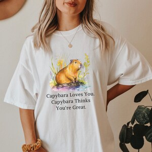 May include: White t-shirt with a watercolor illustration of a capybara in a pond with flowers. The text "Capybara Loves You. Capybara Thinks You're Great." is printed below the image.