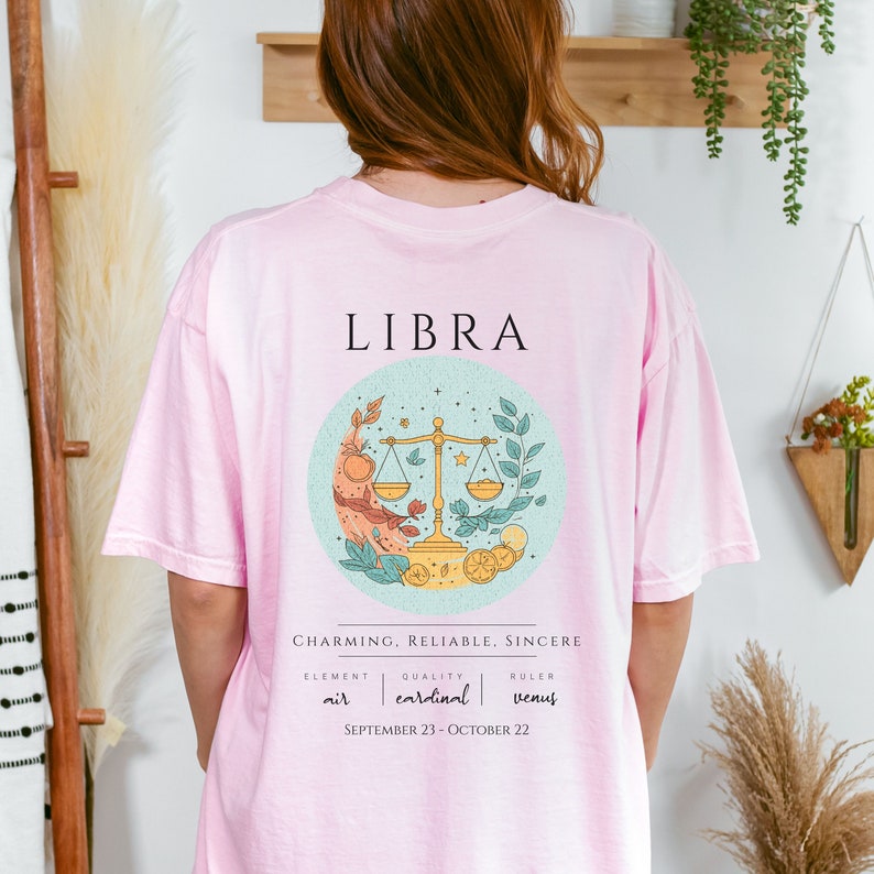 May include: Pink t-shirt with a Libra zodiac sign graphic. The graphic features a light blue circle with a scale, stars, and leaves. The text "LIBRA", "CHARMING, RELIABLE, SINCERE", "ELEMENT air", "QUALITY cardinal", "RULER Venus", and "SEPTEMBER 23-OCTOBER 22" is printed on the shirt.