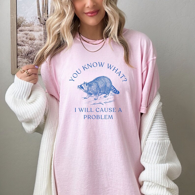 Może przedstawiać: R&oacute;żowy T-shirt z niebieskim nadrukiem przedstawiającym szopa pracza i tekstem "Wiesz co? Sprawię problem."