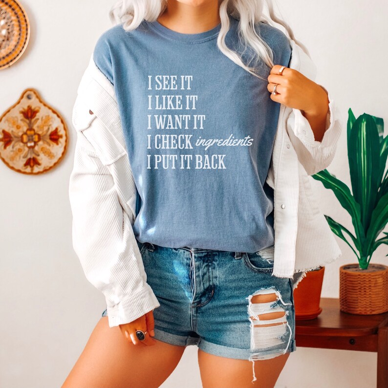 Op de afbeelding: Een lichtblauw T-shirt met een witte afbeelding met de tekst "I see it, I like it, I want it, I check ingredients, I put it back".