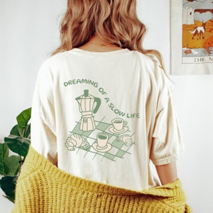 May include: A white t-shirt with a green illustration of a coffee pot, two cups of coffee, and croissants. The text "Dreaming of a slow life" is written in a circle around the illustration.
