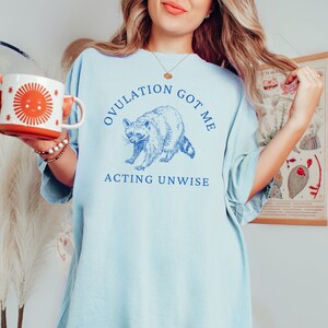 May include: Light blue t-shirt with a raccoon graphic and the text "OVULATION GOT ME ACTING UNWISE".