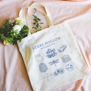 Puede incluir: Una bolsa de lona blanca con texto azul que dice "Stars Hollow * Founded 1779 *" e ilustraciones azules de una cafetera, un cenador, una lib&eacute;lula, un su&eacute;ter, un paraguas, una pila de libros, una pila de panqueques y un muffin de ar&aacute;ndanos.