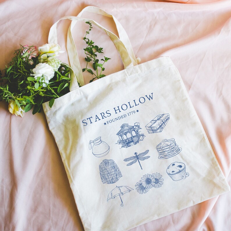 Puede incluir: Una bolsa de lona blanca con texto azul que dice "Stars Hollow * Founded 1779 *" e ilustraciones azules de una cafetera, un cenador, una lib&eacute;lula, un su&eacute;ter, un paraguas, una pila de libros, una pila de panqueques y un muffin de ar&aacute;ndanos.