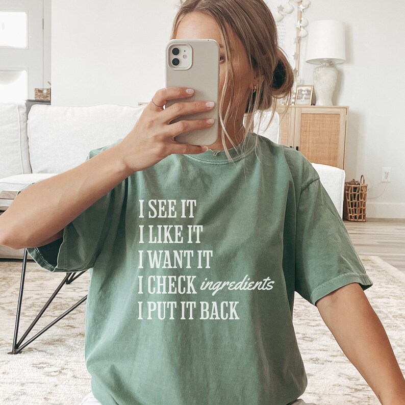 Op de afbeelding: Een groen T-shirt met witte tekst die "I see it, I like it, I want it, I check ingredients, I put it back." zegt.