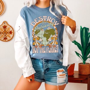 Puede incluir: Una camiseta azul con un gr&aacute;fico de una tierra en forma de coraz&oacute;n con setas y el texto "Destroy the Patriarchy Not the Planet".