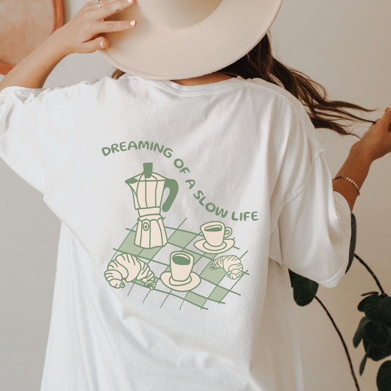 May include: White t-shirt with a green illustration of a coffee pot, two cups of coffee, and croissants. The text "Dreaming of a slow life" is written in green above the illustration.