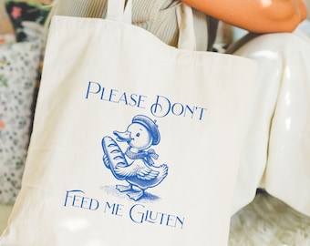 May include: White canvas tote bag with blue text and a cartoon duck holding a baguette. The text reads "Please Don't Feed Me Gluten".