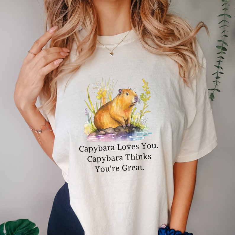 May include: A white t-shirt with a watercolor illustration of a capybara sitting on a rock surrounded by green plants. The text "Capybara Loves You. Capybara Thinks You're Great." is printed below the illustration.