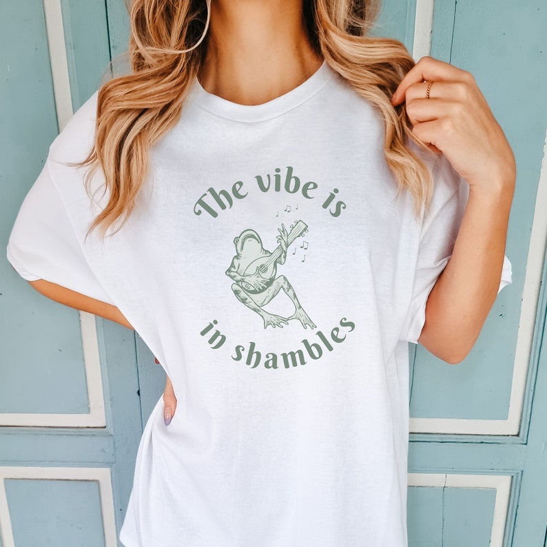 Op de afbeelding: Wit T-shirt met een groene illustratie van een kikker die een snaarinstrument bespeelt. De tekst "The vibe is in shambles" staat boven en onder de kikker in groene kleur.