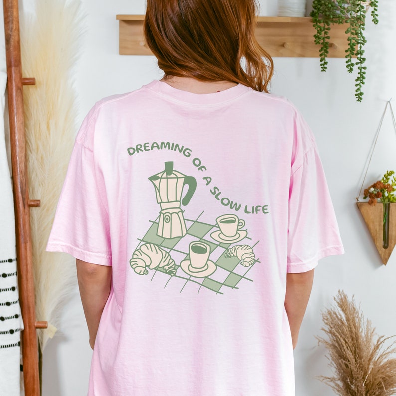 May include: A pink t-shirt with a green graphic of a coffee pot, two cups of coffee, and croissants on a checkered surface. The text "DREAMING OF A SLOW LIFE" is printed in a circle around the graphic.