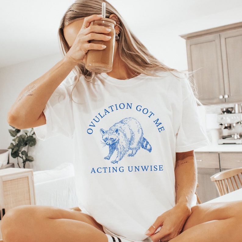 May include: White t-shirt with a blue graphic of a raccoon and the text "OVULATION GOT ME ACTING UNWISE".