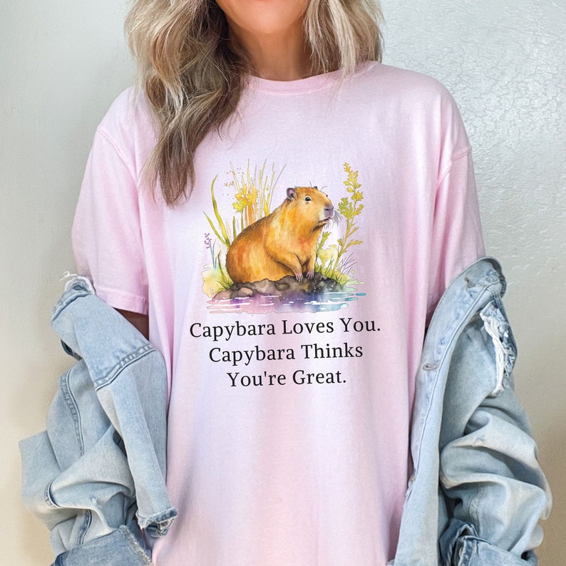 May include: A pink t-shirt with a watercolor illustration of a capybara sitting in a grassy area. The text "Capybara Loves You. Capybara Thinks You're Great." is printed below the illustration.