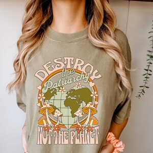 Puede incluir: Una camiseta verde con un dise&ntilde;o gr&aacute;fico retro. El gr&aacute;fico presenta una tierra en forma de coraz&oacute;n con hongos y flores alrededor. El texto "Destroy the Patriarchy Not the Planet" est&aacute; escrito en una fuente retro colorida.
