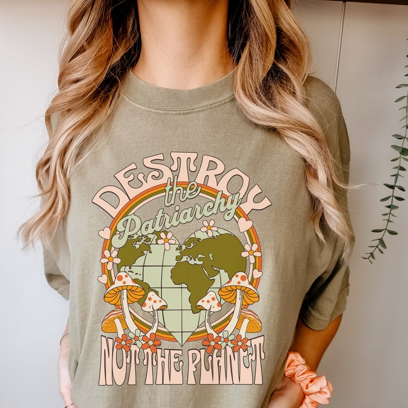Puede incluir: Una camiseta verde con un dise&ntilde;o gr&aacute;fico retro. El gr&aacute;fico presenta una tierra en forma de coraz&oacute;n con hongos y flores alrededor. El texto "Destroy the Patriarchy Not the Planet" est&aacute; escrito en una fuente retro colorida.