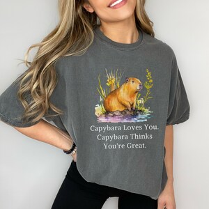 May include: A dark gray t-shirt with a watercolor illustration of a capybara sitting in a pond. The text "Capybara Loves You. Capybara Thinks You're Great." is printed below the image.