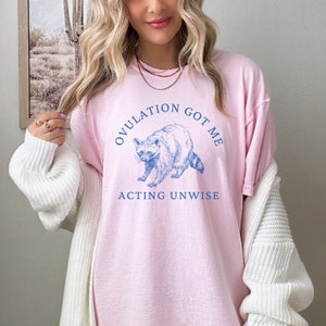 May include: Pink t-shirt with a blue illustration of a raccoon and the text "OVULATION GOT ME ACTING UNWISE".