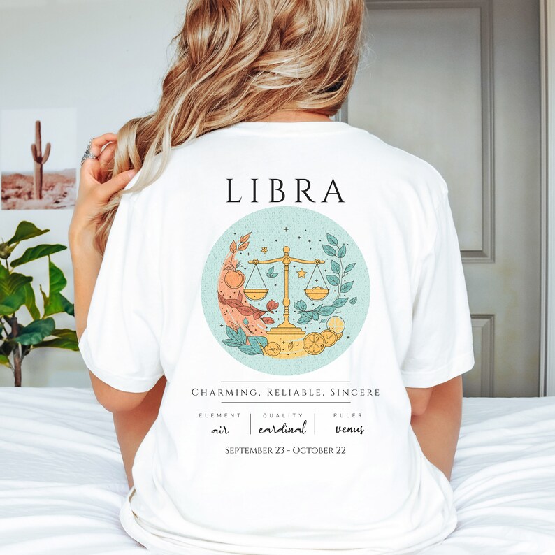 May include: White t-shirt with a graphic of the Libra zodiac sign. The graphic features a teal circle with a gold scale, leaves, and lemons. The text "LIBRA" is at the top of the shirt and the text "CHARMING, RELIABLE, SINCERE" is below the graphic. The text "ELEMENT air, QUALITY cardinal, RULER venus" and "SEPTEMBER 23 - OCTOBER 22" are below the text.