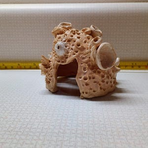 May include: A coral-like, beige ceramic sculpture with a porous texture and irregular shapes. The sculpture has multiple openings and protrusions, resembling a natural sea formation. A yellow measuring tape is in the background.