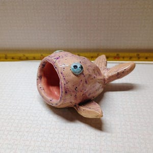 May include: A ceramic fish-shaped container with a wide open mouth, fins, and tail. The fish is light pink with purple speckles and blue eyes. The inside of the mouth is a darker pink.