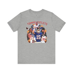 May include: A gray t-shirt with a graphic print of a football player wearing a blue jersey with the number 15. The text "TOMMY CUTLETS" is printed in red above the graphic.