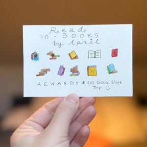 Può includere: Un biglietto disegnato a mano con il testo "Read 10 Books by April" e illustrazioni di libri. Il biglietto include anche il testo "Reward: 100$ Book Store Trip."