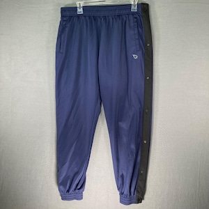 Può includere: Pantaloni sportivi blu navy con una striscia nera sul lato con chiusure a scatto. I pantaloni hanno vita e polsini elasticizzati. Un piccolo logo è visibile sulla gamba sinistra.