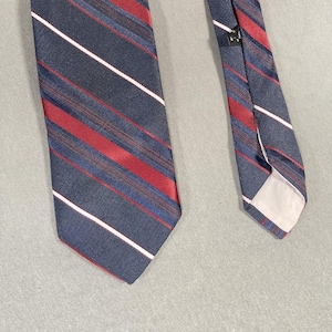 Puede incluir: Una corbata azul marino con rayas diagonales rojas, blancas y azules. La corbata está hecha de una tela texturizada y está en ángulo sobre una superficie gris claro. Un accesorio clásico.