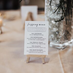 May include: A white card with black text on an easel, titled "Kissing Menu". The menu lists different ways for guests to show their love for the newlyweds, such as "The Songbird", "The Entertainer", "The French Toast", "The Example", and "The Heartwarmer".