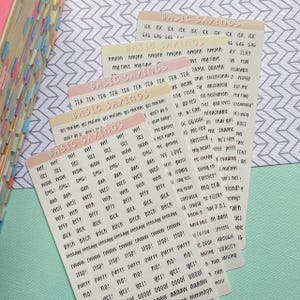 May include: A stack of sticker sheets with the words "BASIC SAYINGS" printed at the top. The stickers have various phrases and words in a handwritten font, including "OMG!", "WOW", and "STOP!" The stickers are in shades of white, pink, and beige.