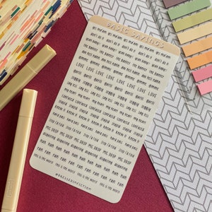 May include: A sticker sheet with the words "BASIC SAYINGS" at the top. The sheet is filled with various phrases in a handwritten font. Two beige highlighters and a ruler are also visible, along with a maroon surface and patterned paper.