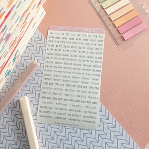 May include: A sticker sheet with the text "BASIC SAYINGS" in black font. The sheet is filled with phrases like "weak," "obsessed," and "the audacity." The background is a mix of pink and patterned paper, with markers and a ruler.