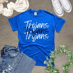 May include: A royal blue t-shirt with the word "Trojans" in a layered design. The shirt is paired with blue jeans and white sneakers. Decorative elements include white flowers and greenery.