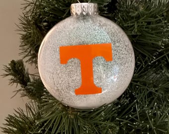May include: A white Christmas ornament with a glittery finish and an orange "T" on the front.