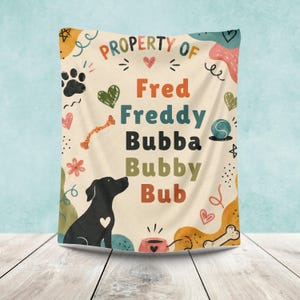 Puede incluir: Una manta de color crema con el texto Property of Fred, Freddy, Bubba, Bubby, Bub en varios colores. El diseño incluye la silueta de un perro negro, huellas, corazones, un hueso, una pelota y formas abstractas en naranja, verde y azul.
