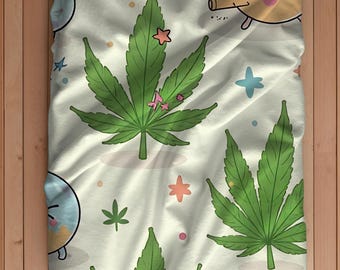 May include: A white blanket with a repeating pattern of large green cannabis leaves, cartoon stars, and other whimsical shapes. The blanket has a soft, inviting appearance, perfect for a cozy setting.