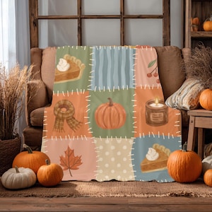 May include: A soft throw blanket featuring a patchwork design with illustrations of pumpkin pie slices, a plaid scarf, a central orange pumpkin, a lit candle, a maple leaf, and berries, all accented by white stitched lines on a muted colour palette.