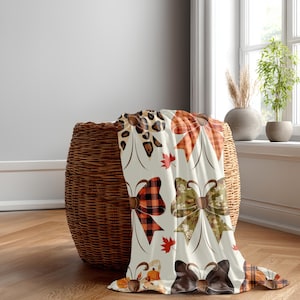 May include: A cream-colored throw blanket featuring a repeating pattern of various bows and small red maple leaves. The bows include designs such as leopard print, orange plaid, red and black buffalo check, and olive green floral patterns.