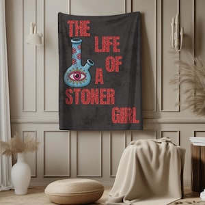 May include: A black tapestry with the text "THE LIFE OF A STONER GIRL" in red glitter lettering. A blue bong with pink flowers and an eye design is on the left side of the tapestry.
