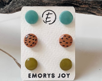 May include: A set of six colorful stud earrings on a white card with the text "Emory's Joy". The earrings are in three different colors: teal, brown with black spots, and olive green.