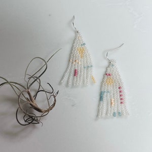 May include: A pair of white beaded tassel earrings with colorful accents. The earrings are made with small, white beads and feature pops of pink, yellow, and blue.