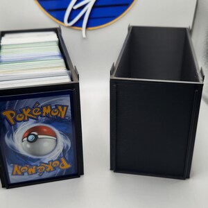 Può includere: Due scatole rettangolari in plastica nera per conservare carte collezionabili. Una scatola è riempita con una pila di carte, con una carta Pokémon blu sul davanti che mostra il logo Poké Ball e del testo. L'altra scatola è vuota e aperta.